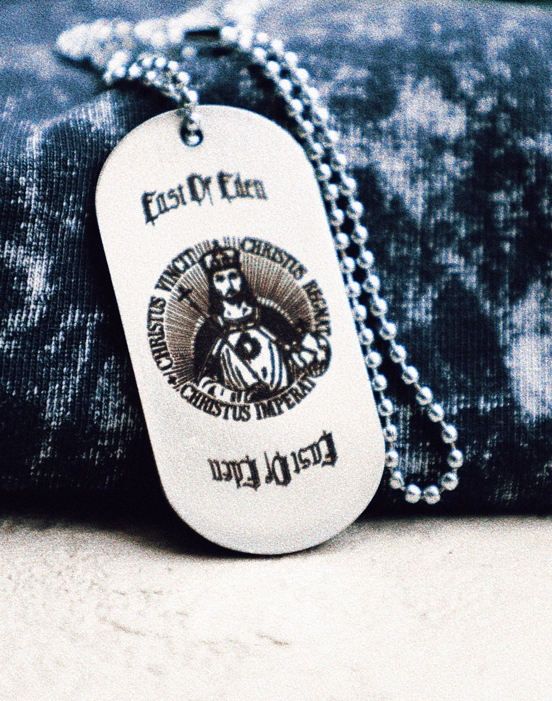 Close-up of the back side of the SOL (Sovereign Order of the Lamb) metal dog tag, featuring gothic text reading “East of Eden” above an illustration of Christ the King surrounded by the Latin phrase “Christus Vincit, Christus Regnat, Christus Imperat,” resting on a textured acid-washed fabric. A symbolic accessory from the grunge Indian Christian streetwear collection.