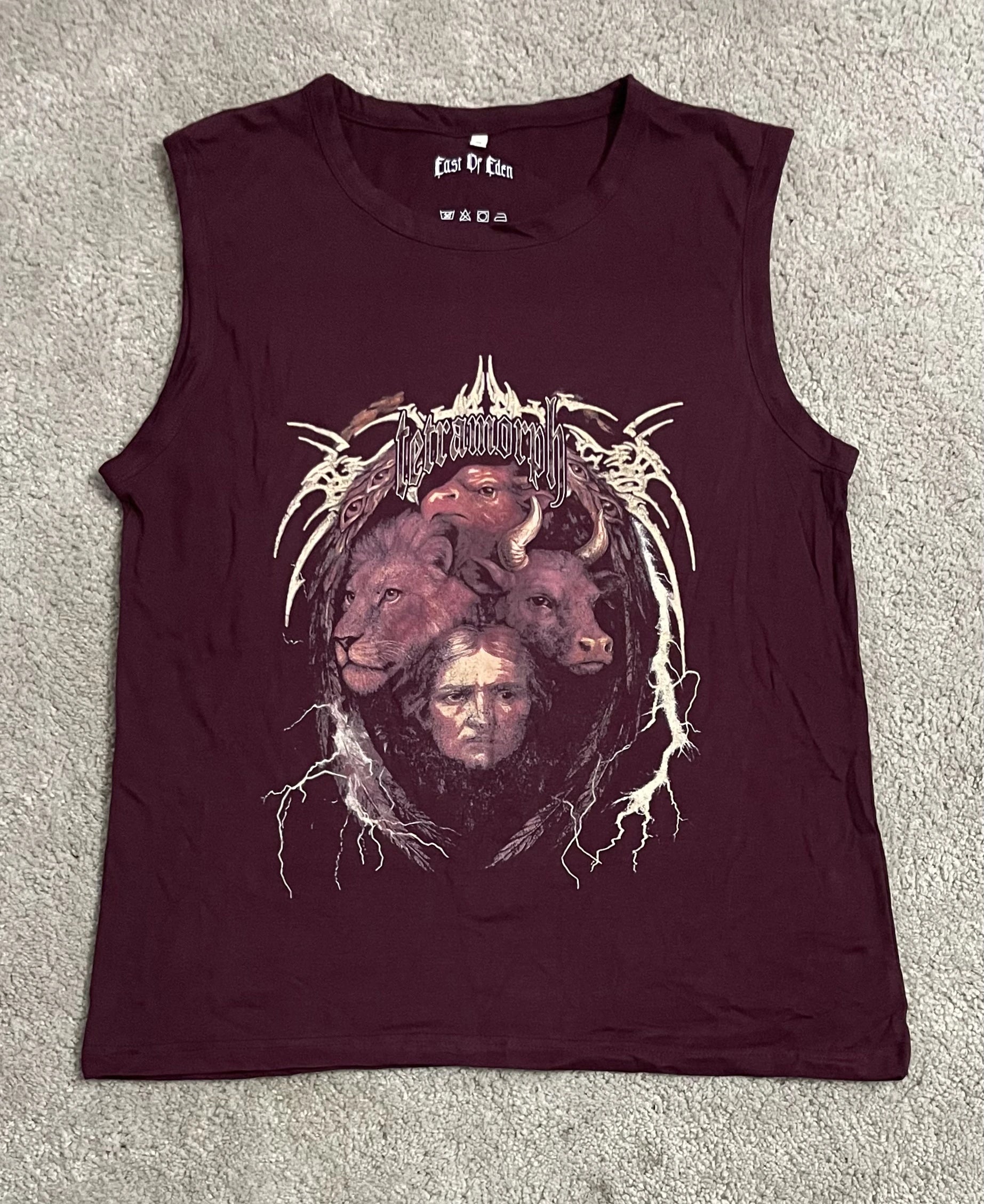 Maroon sleeveless tank top featuring the Tetramorph graphic from Revelation, depicting a lion, ox, eagle, and man framed by lightning-like accents and gothic lettering, laid flat on a textured surface. A striking piece from the grunge Indian Christian streetwear collection.