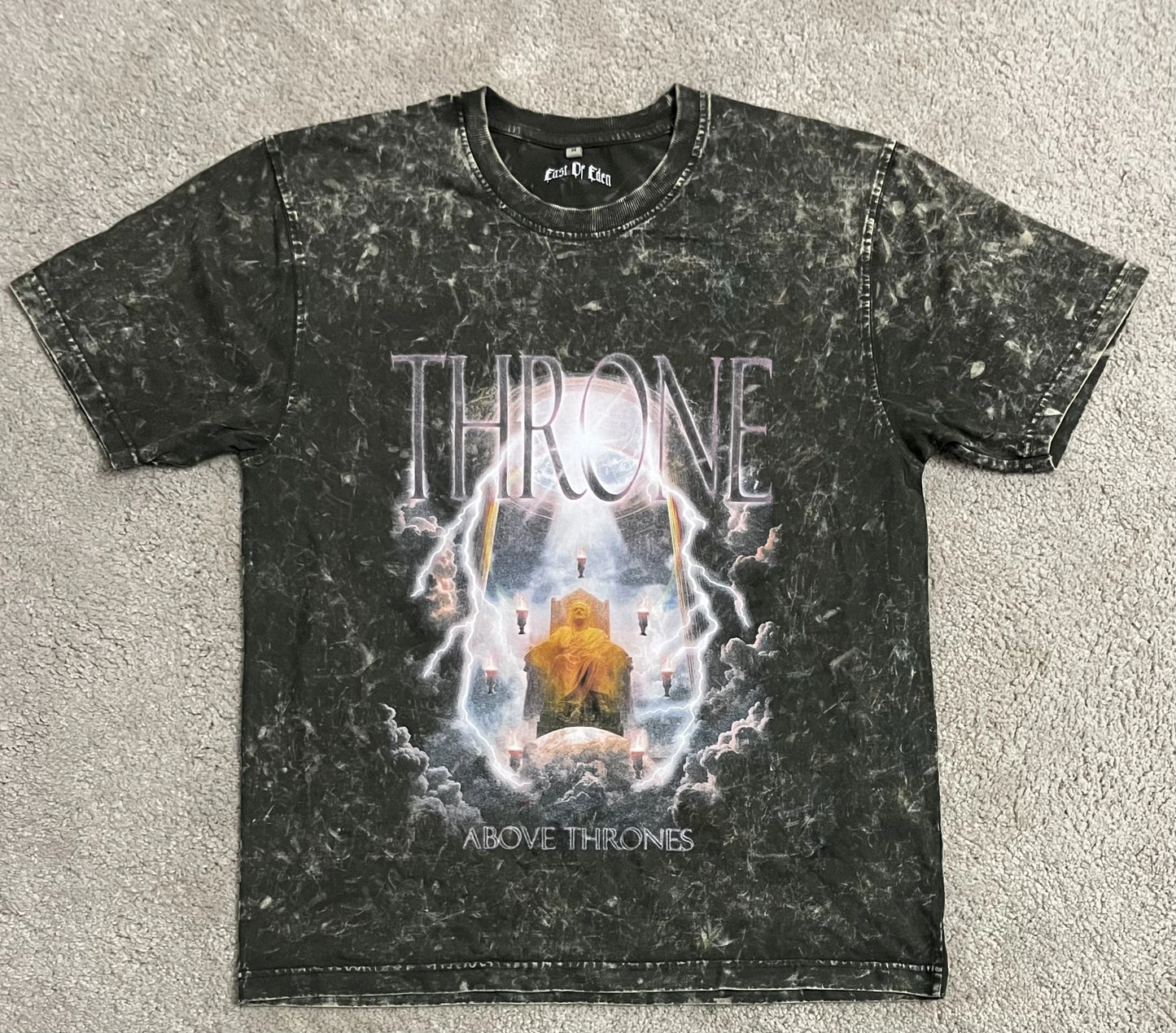 Olive green acid-washed t-shirt featuring the “Throne Above Thrones” graphic inspired by Revelation, showing a radiant enthroned figure surrounded by lightning and clouds, laid flat on a gray surface. A striking piece from the grunge Indian Christian streetwear collection.