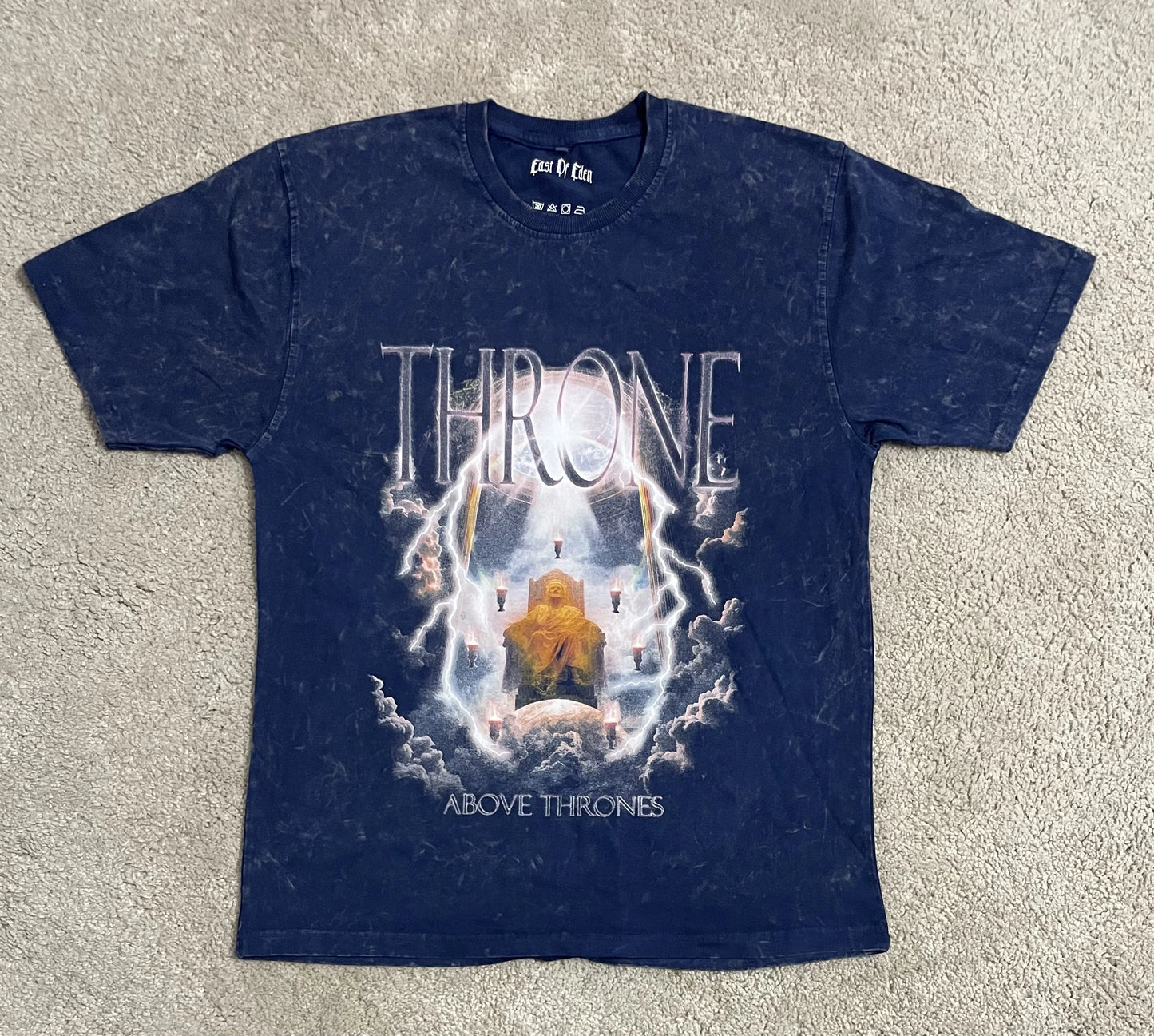 Navy blue acid-washed t-shirt featuring the “Throne Above Thrones” graphic inspired by Revelation, depicting a glowing enthroned figure surrounded by lightning and clouds, laid flat on a gray surface. A bold design from the grunge Indian Christian streetwear collection.