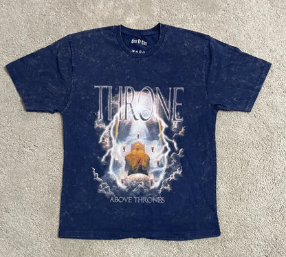 Navy blue acid-washed t-shirt featuring the “Throne Above Thrones” graphic inspired by Revelation, depicting a glowing enthroned figure surrounded by lightning and clouds, laid flat on a gray surface. A bold design from the grunge Indian Christian streetwear collection.