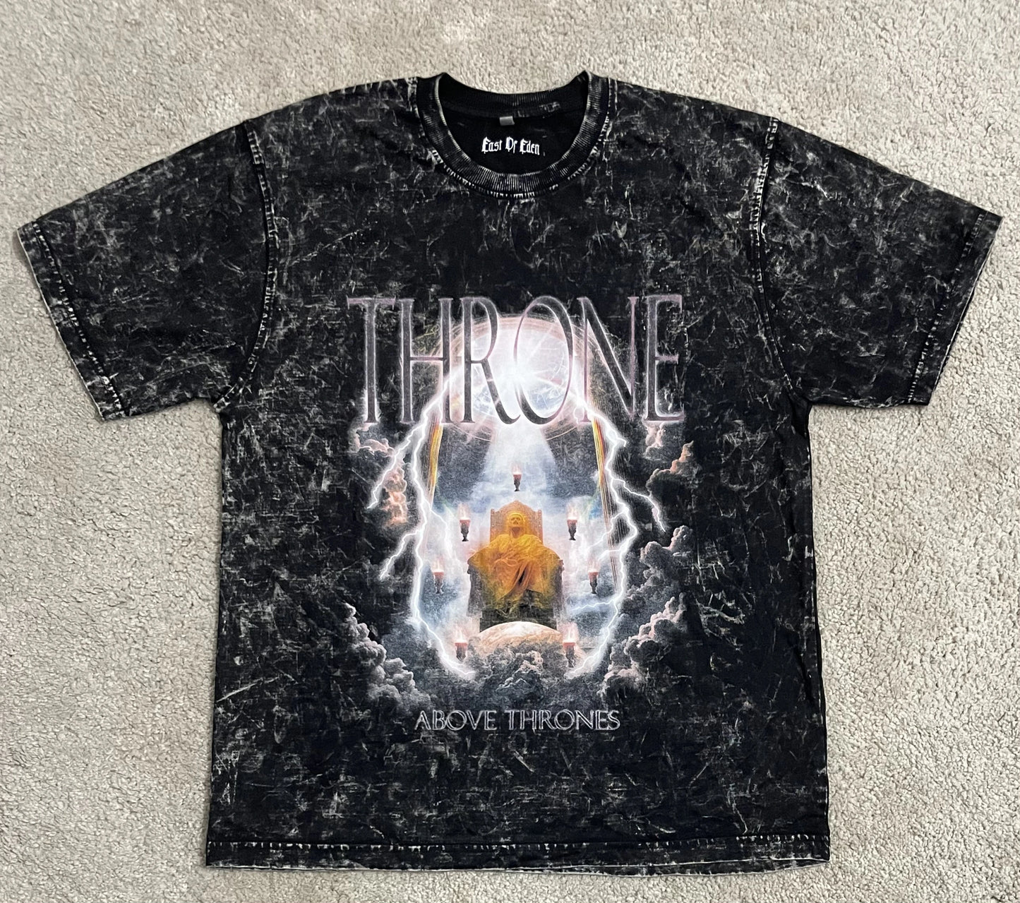 Black acid-washed t-shirt featuring the “Throne Above Thrones” graphic inspired by Revelation, with a radiant enthroned figure surrounded by lightning and clouds, laid flat on a neutral background. A powerful piece from the grunge Indian Christian streetwear collection.