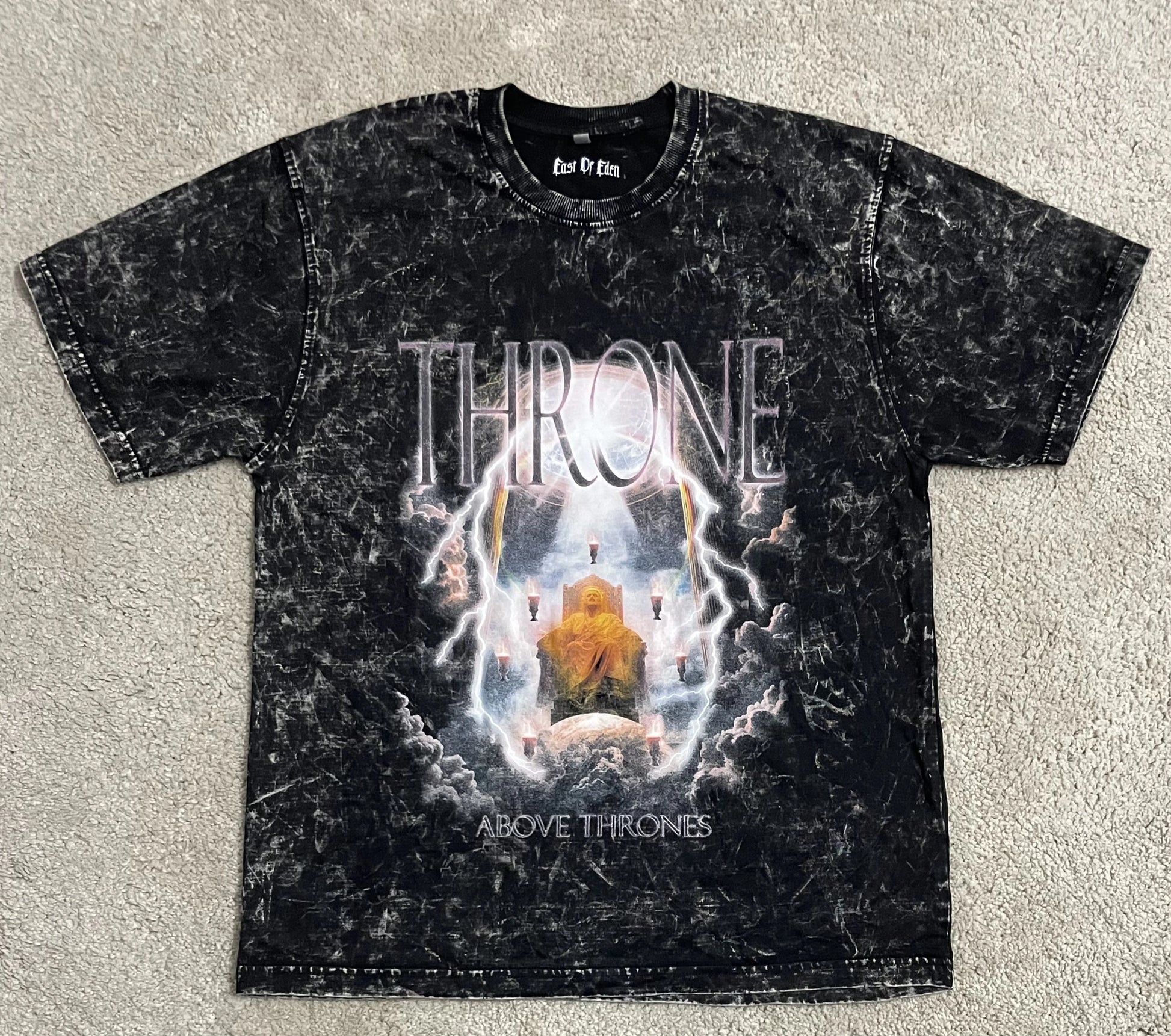 Black acid-washed t-shirt featuring the “Throne Above Thrones” graphic inspired by Revelation, with a radiant enthroned figure surrounded by lightning and clouds, laid flat on a neutral background. A powerful piece from the grunge Indian Christian streetwear collection.