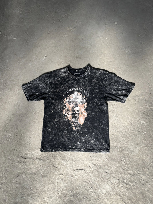 Black acid-washed t-shirt featuring the “Deathless” graphic with a skull and ornate cross referencing “1 Corinthians 15:55,” laid flat on a gray concrete surface. The first drop from the grunge Indian Christian streetwear collection.