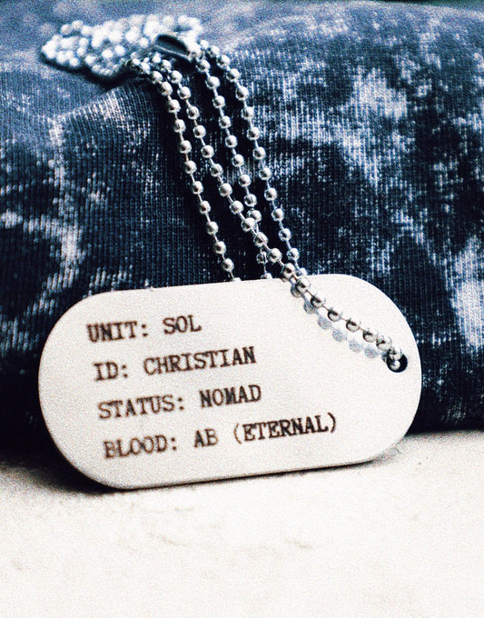 Close-up of the SOL (Sovereign Order of the Lamb) metal dog tag on a textured acid-washed fabric, engraved with the text “UNIT: SOL, ID: CHRISTIAN, STATUS: NOMAD, BLOOD: AB (ETERNAL),” symbolizing faith and identity in the grunge Indian Christian streetwear collection.