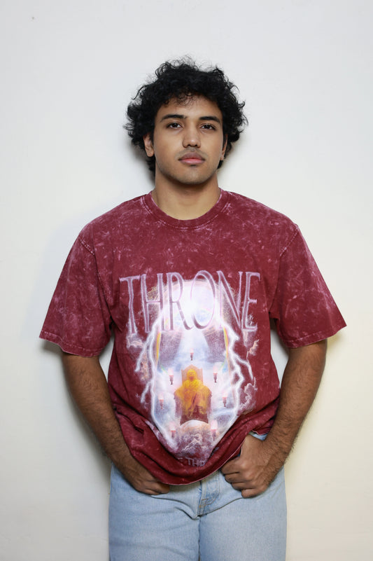 Model wearing a maroon acid-washed t-shirt featuring the “Throne Above Thrones” graphic from Revelation, with a radiant figure seated on a heavenly throne surrounded by lightning and seven flames. A bold piece from the grunge Indian Christian streetwear collection.